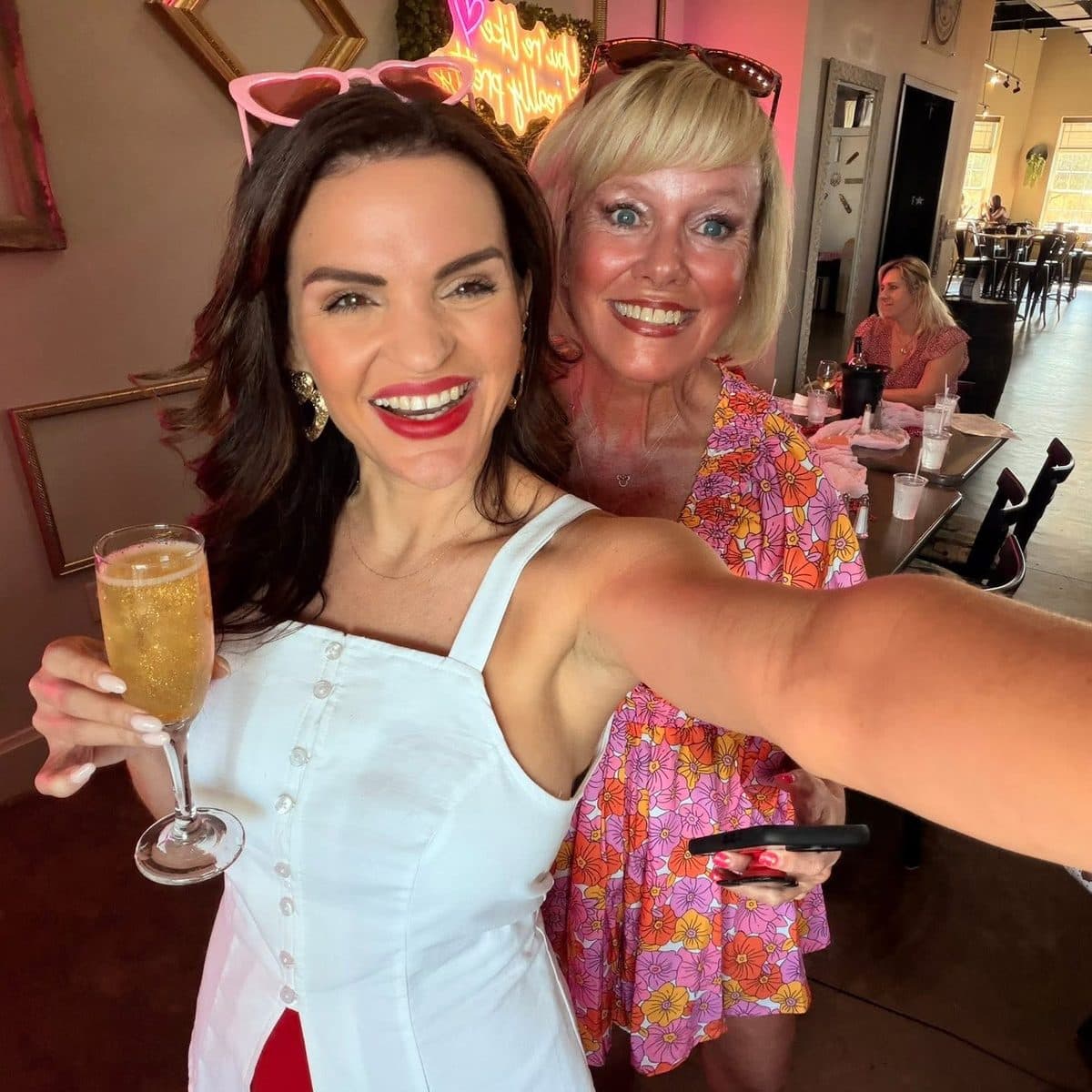 Luana and a guest smiling in a selfie at the Galentine's brunch, with a pink neon sign behind them