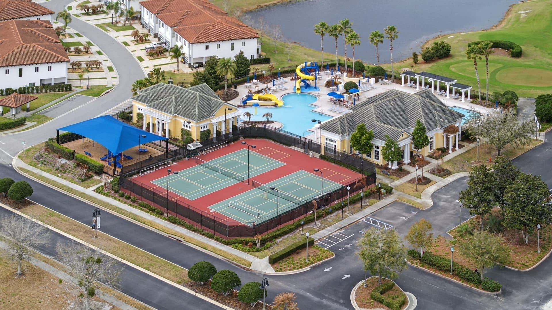 Aerial view of two yellow amenity buildings in Providence with a pool and yellow water slides, fenced tennis courts, and a shaded playground, with a lake and the golf course behind